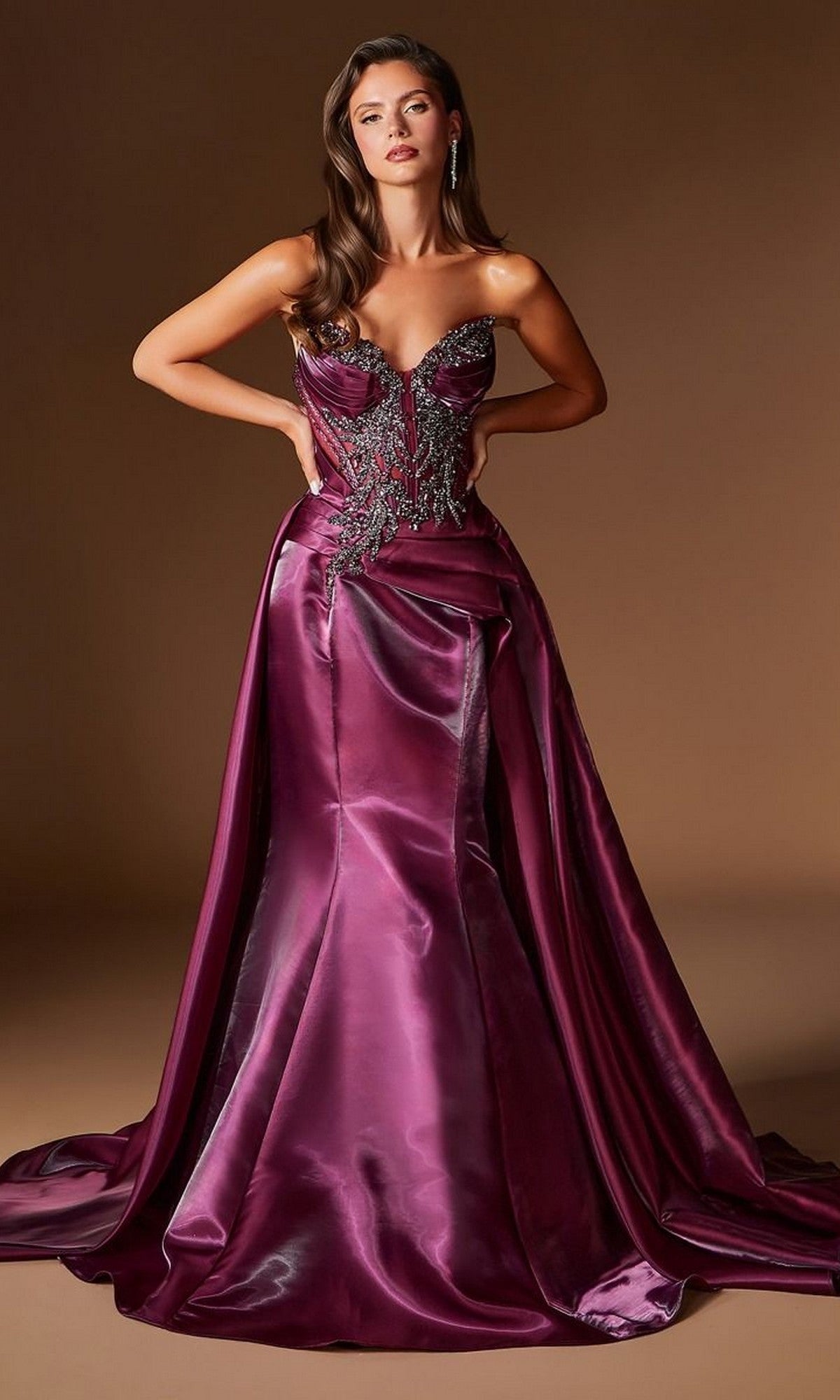 A woman stands against a plain brown background wearing the Ladivine CDS568 long prom dress in Plum Noir, featuring a strapless, floor-length satin design with hand-beaded embellishments on the bodice.
